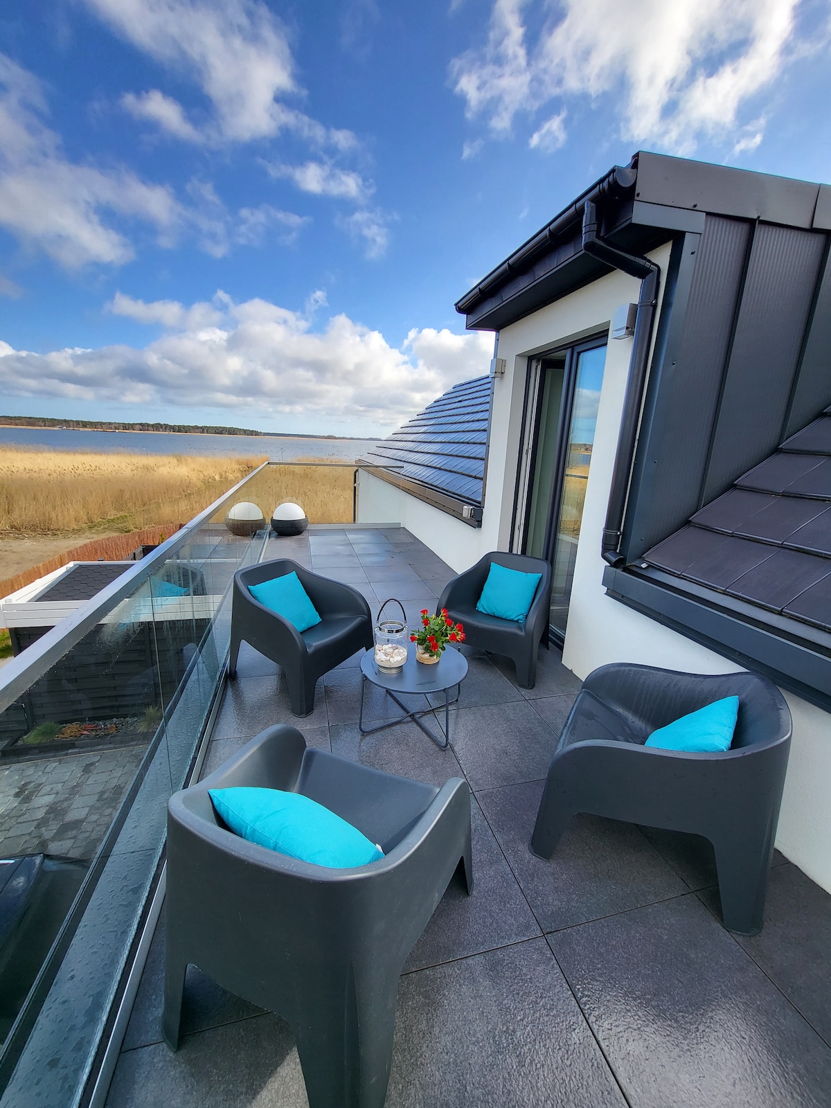 A serene terrace provides a panoramic view of the landscape, featuring four modern chairs with bright blue cushions arranged around a small table. The space is enhanced by a glass railing, allowing for an unobstructed view of the surrounding area.