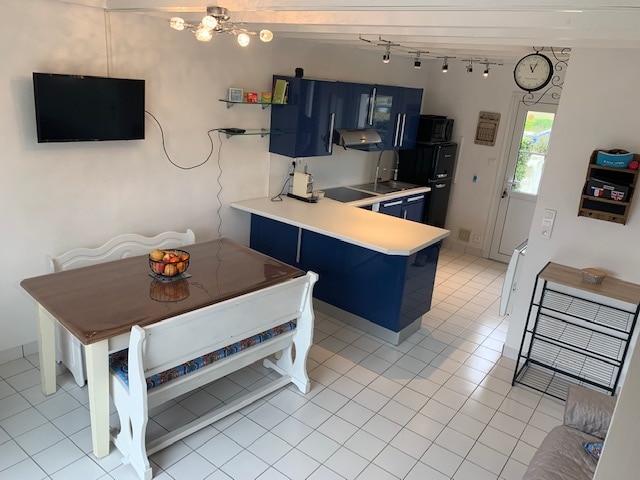 A modern, open-plan kitchen and dining area is presented, featuring sleek blue cabinetry and a dining table with a fruit bowl at its center. White tiled flooring complements the clean design, with a wall-mounted TV and clock adding functional touches.