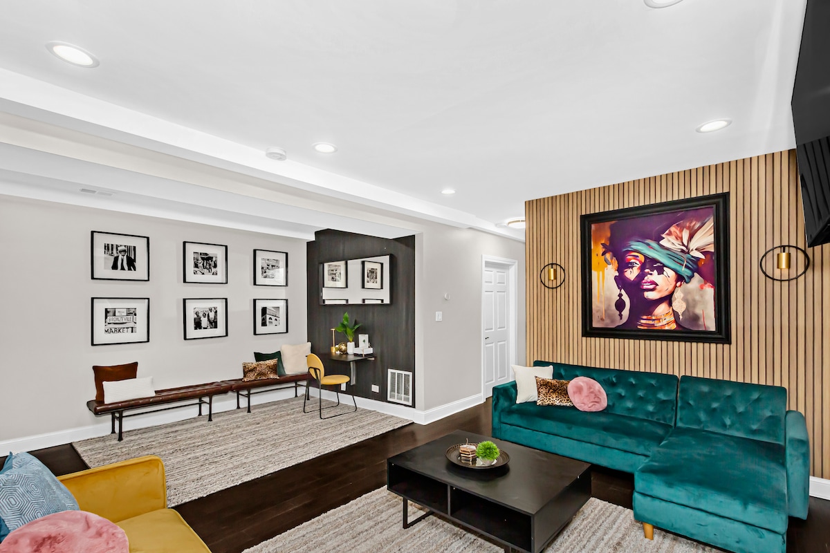 A spacious living area is showcased, featuring a vibrant teal sofa and a classic wall-mounted television. A gallery of black and white framed artworks decorates one wall, while a wooden bench and green decorative plants provide additional seating and visual interest.
