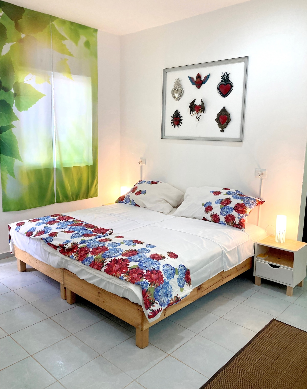A cozy sleeping area features a spacious bed adorned with floral bedding, complemented by soft lighting from bedside lamps. A wall adorned with decorative art adds visual interest, while natural light filters through a large window, enhancing the serene atmosphere.