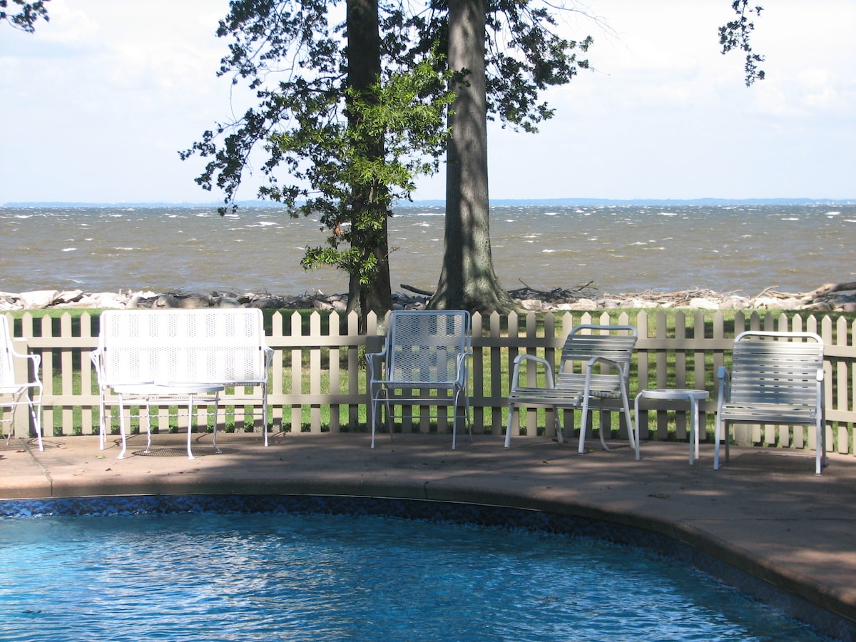 A swimming pool is framed by a stone patio and surrounded by several white chairs. Beyond the pool, a view of the Chesapeake Bay is visible, lined by trees and a picket fence. The sky is partly cloudy, suggesting a breezy atmosphere.