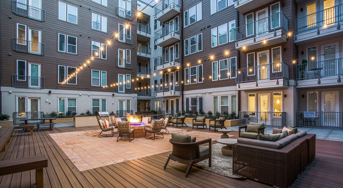 An outdoor courtyard area features a central fire pit surrounded by comfortable seating arrangements. String lights hang overhead, providing soft illumination. Multiple stories of residential apartments encircle the space, with large windows visible in each unit.