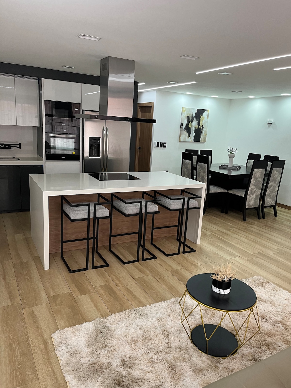 A modern kitchen is featured, showcasing a white quartz island with seating for three. Stainless steel appliances are integrated, including a refrigerator and oven. A dining area accommodates six, while soft lighting highlights the sleek finishes and clean design of the space.