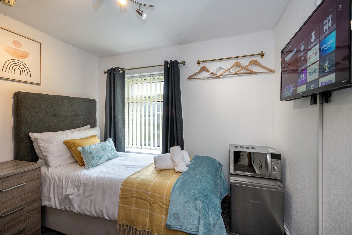 A cozy single room features a single bed with white bedding and a decorative teal throw. A television is mounted on the wall, and a microwave is placed next to a small fridge. Light enters through a window with vertical blinds, enhancing the room's welcoming feel.