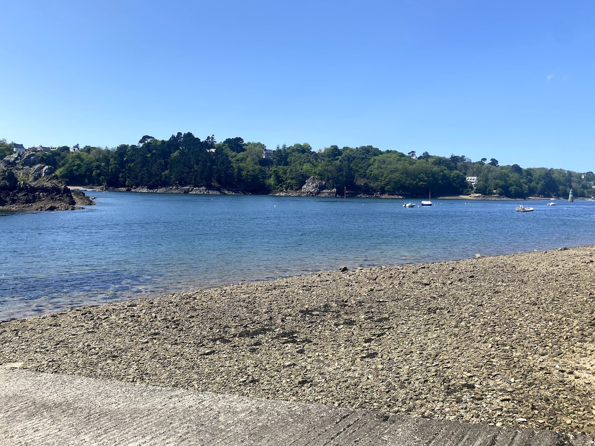 A calm waterfront scene is showcased, with clear blue water gently lapping against a rocky shore. Lush greenery outlines the opposite bank, while small boats can be seen on the water. The sky is clear and bright, adding to the serene atmosphere.