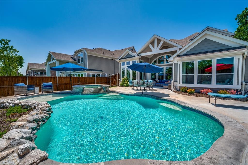 A spacious backyard features a large, inviting pool with a tanning ledge and built-in hot tub. Umbrellas provide shade over seating areas, while a water feature softly cascades. Both the house and pool area are surrounded by a tasteful fence, ensuring privacy.