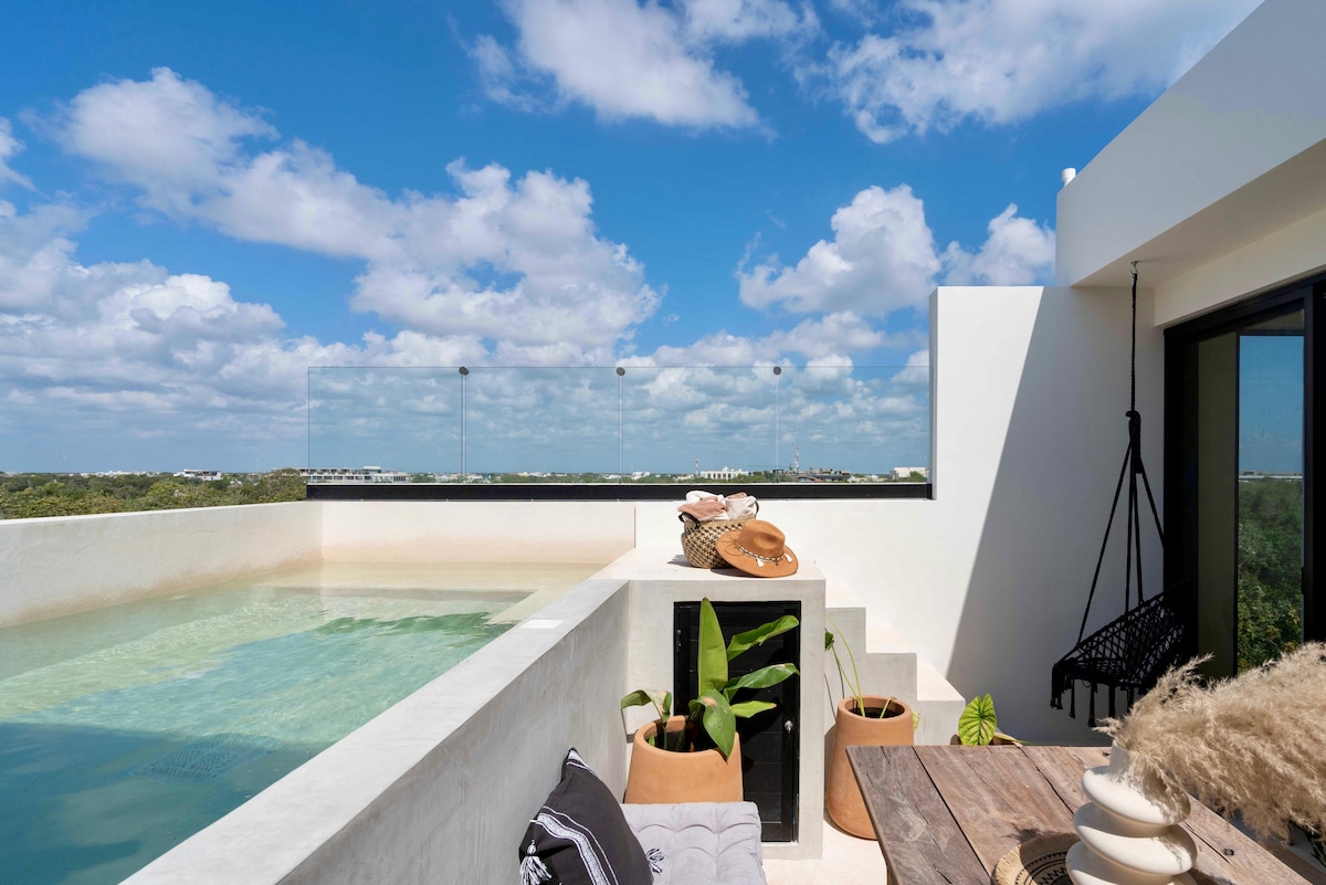 An outdoor space is presented with a private pool surrounded by smooth, light-colored walls. A cozy seating area is furnished with cushions and a decorative throw. Potted plants add a touch of greenery, while an expansive sky with scattered clouds provides a bright backdrop.