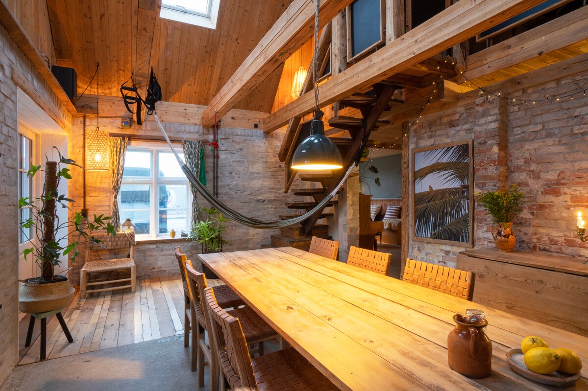 A spacious dining area features a large wooden table surrounded by woven chairs. A hammock is suspended nearby, adding relaxation to the design. Warm light from hanging fixtures illuminates the space, highlighting the natural wood beams and stone walls, while greenery softly decorates the room.