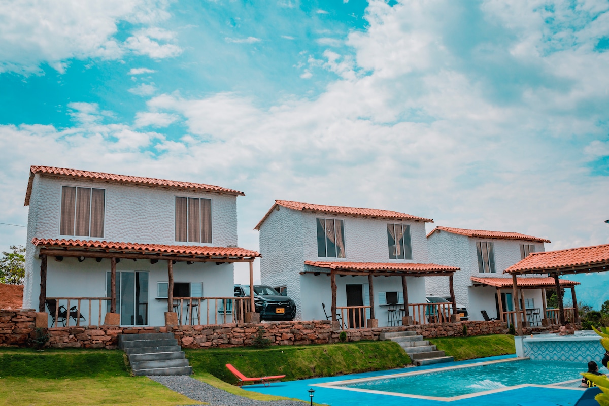 Three charming cabins are positioned alongside a pool, featuring distinctive terracotta roofs and white exteriors. A well-maintained lawn surrounds the area, with stone pathways leading to the cabins. The sky above is clear with scattered clouds, creating a serene atmosphere.