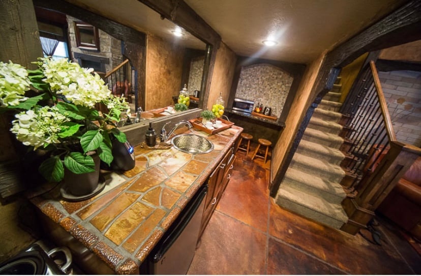A rustic kitchen area features a stone countertop with a stainless steel sink and modern appliances. Potted green plants and fresh flowers are displayed, adding a touch of nature. A staircase leads to the upper level, while warm lighting enhances the inviting ambiance.