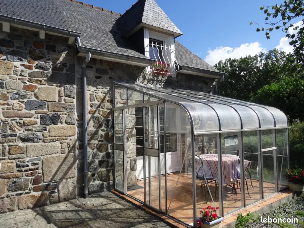 A glass-enclosed veranda features a warm wooden floor and a table set for two, with vibrant flowers placed along the edges. The stone exterior of the farmhouse is visible, complemented by slate roof tiles and a small window above the entrance. Natural light fills the space.