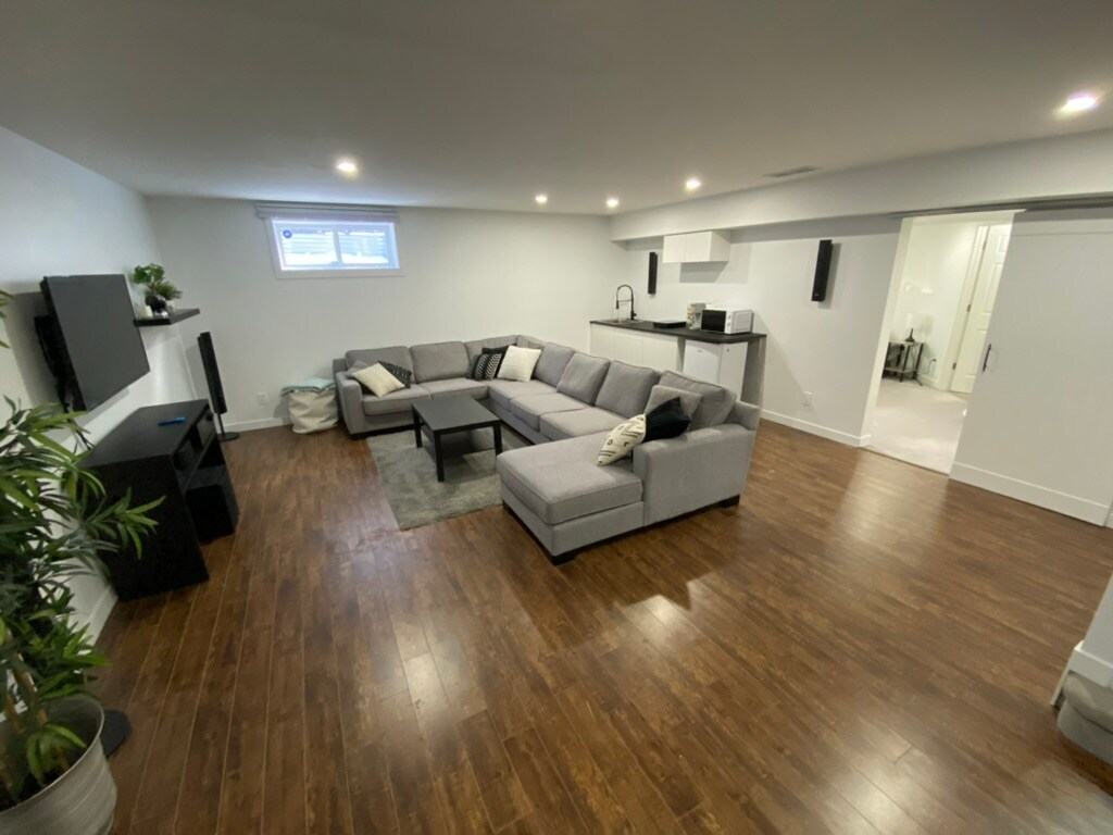 A spacious living area features a large sectional sofa arranged around a coffee table, with a television mounted on the wall. Soft lighting illuminates the space, complemented by sleek wooden flooring. A kitchenette area is visible in the background, along with a doorway leading to an ensuite bathroom.