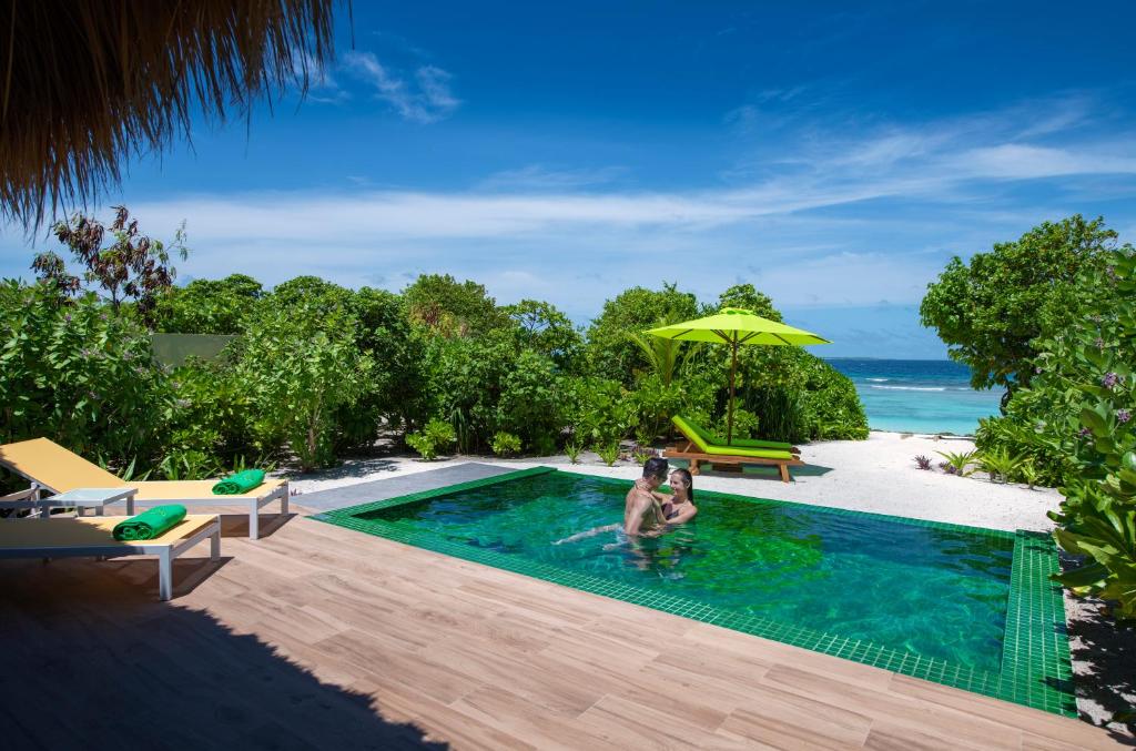 A private pool is set within a tropical landscape, surrounded by lush greenery. Two sun loungers in yellow and green are positioned nearby, while a bright green umbrella provides shade. The ocean is visible beyond the pool, under a clear blue sky.