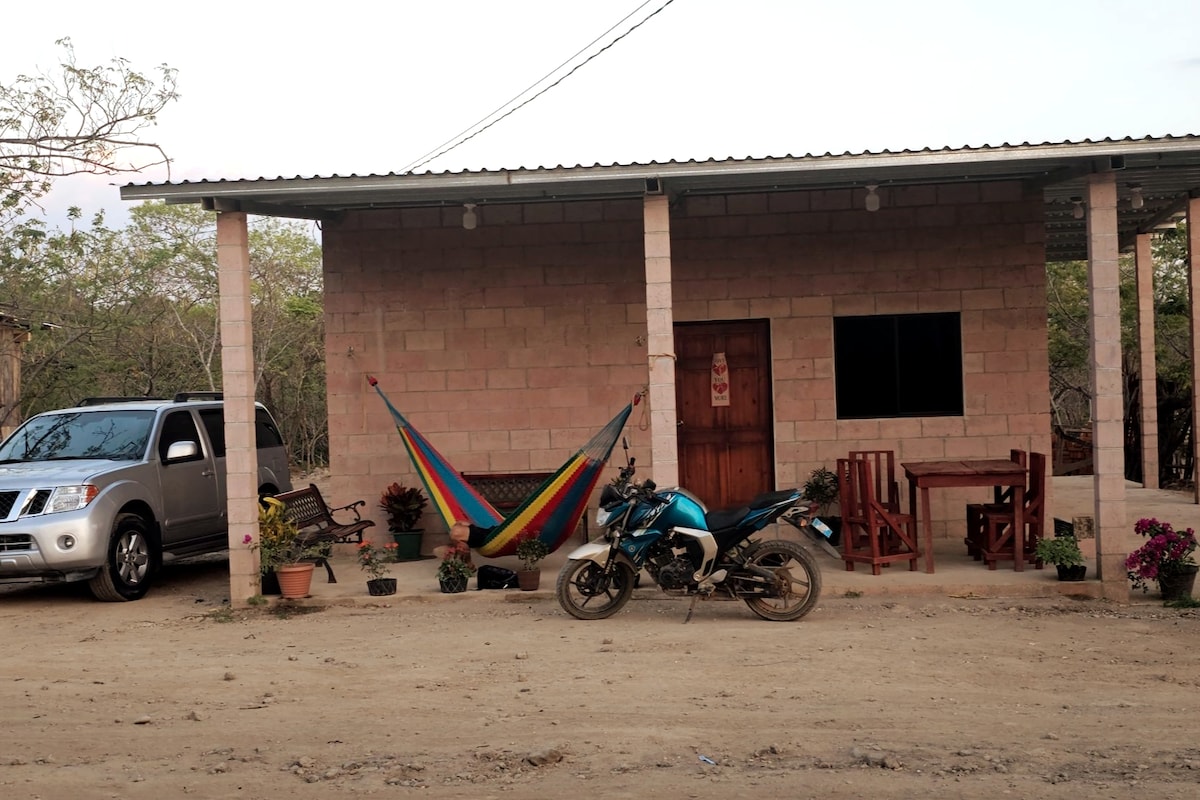 A modest countryside house is presented with a light-colored stone facade. In front, a colorful hammock is suspended between two posts. A silver vehicle and a blue motorcycle are parked nearby, with a rustic wooden table and chairs positioned on the dirt surface, surrounded by potted plants.
