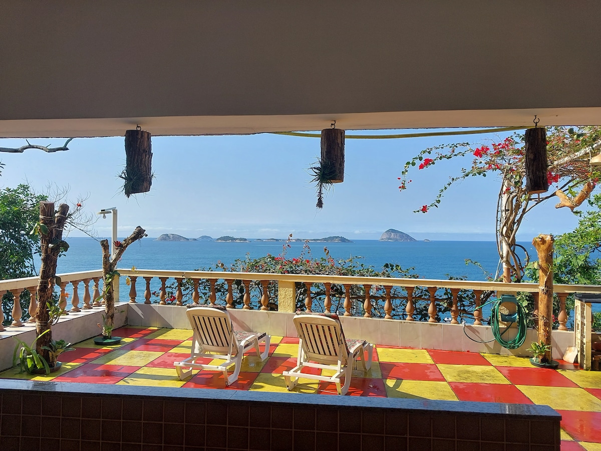 A spacious terrace features two lounge chairs overlooking a vibrant blue ocean. Colorful tiles in red and yellow cover the floor, while flowers and greenery embellish the railings. The scene is framed by distant islands on the horizon under a clear sky.