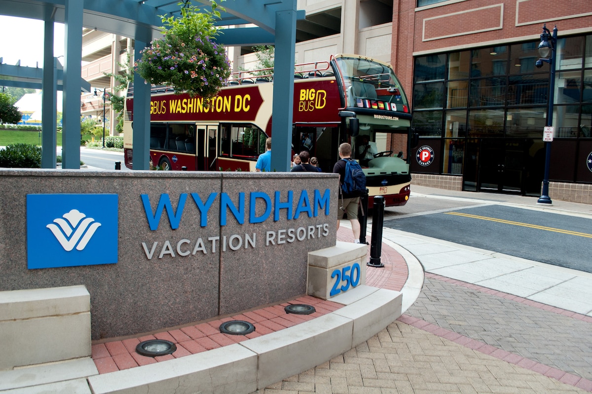 The entrance to Wyndham Vacation Resorts is depicted, showcasing a concrete sign with the Wyndham logo prominently featured. A double-decker bus labeled 'Big Bus Washington DC' is seen parked nearby, while guests prepare to board. The surrounding area displays landscaped greens and urban architecture.