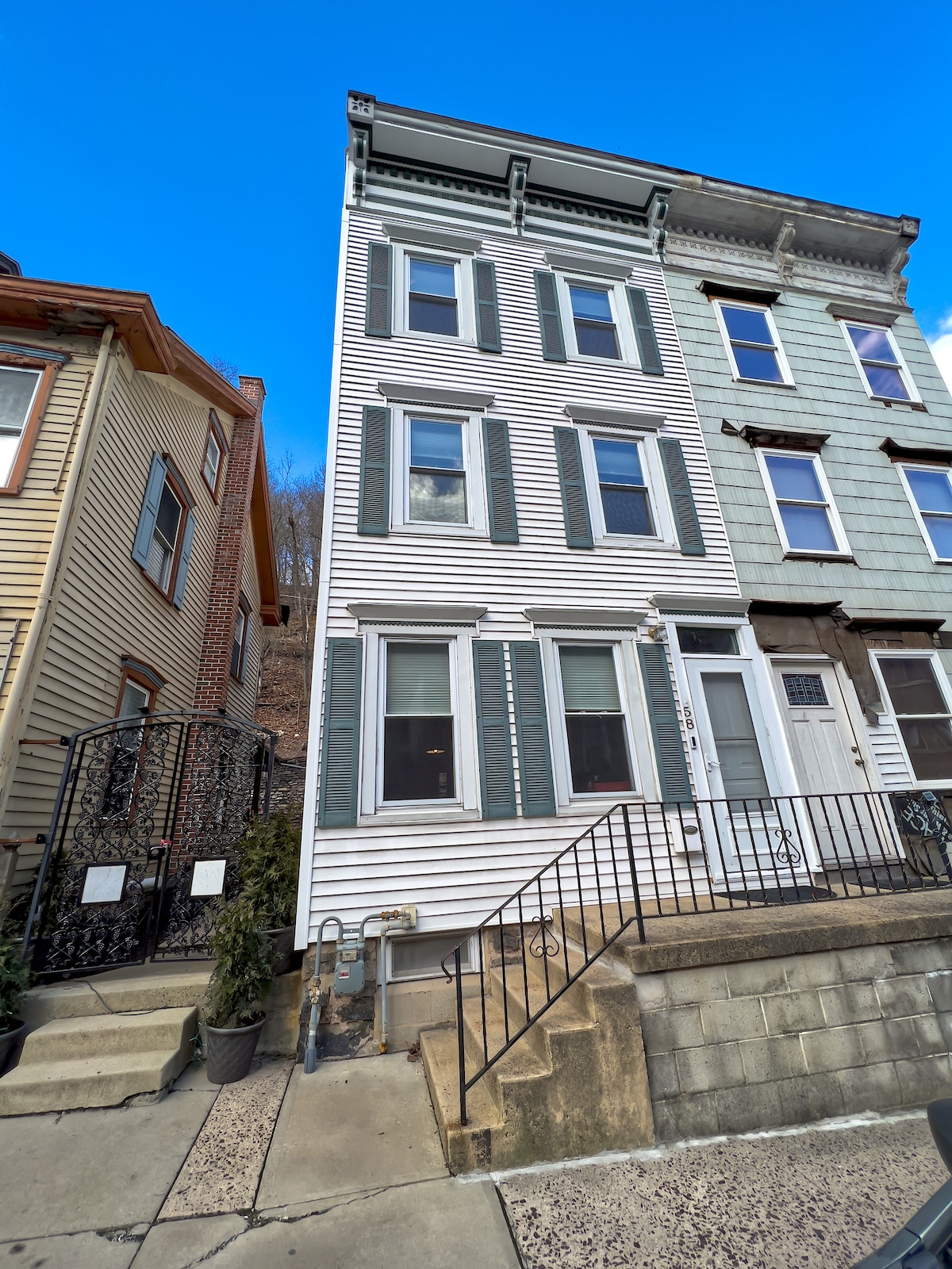 A three-story house with white siding and green shutters is shown. The entrance features a set of steps leading to a door, flanked by large windows on the front facade. A neighboring building is partially visible on the left.
