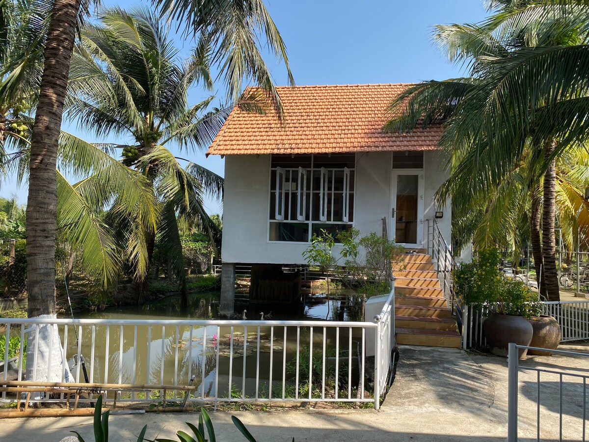 A charming house is positioned on stilts, surrounded by lush palm trees. The exterior features a combination of white walls and a red-tiled roof. A staircase leads to the entrance, with a serene pond visible at the base.