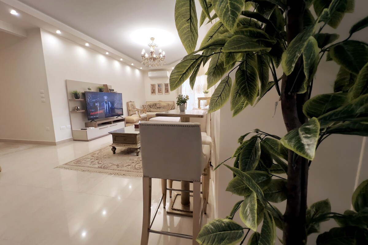 A modern living area is presented, featuring a neutral color palette and elegant furnishings. A comfortable seating arrangement surrounds a flat-screen television, and a decorative rug adds warmth to the space. A green potted plant is positioned to the side, enhancing the inviting ambiance.