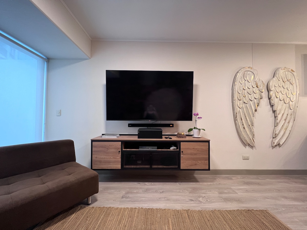 A modern living area features a large flat-screen TV mounted on a wooden media console, which houses storage compartments. A comfortable brown couch provides seating, while decorative wall art in the shape of angel wings adds an artistic touch. Natural light enters through a nearby window.