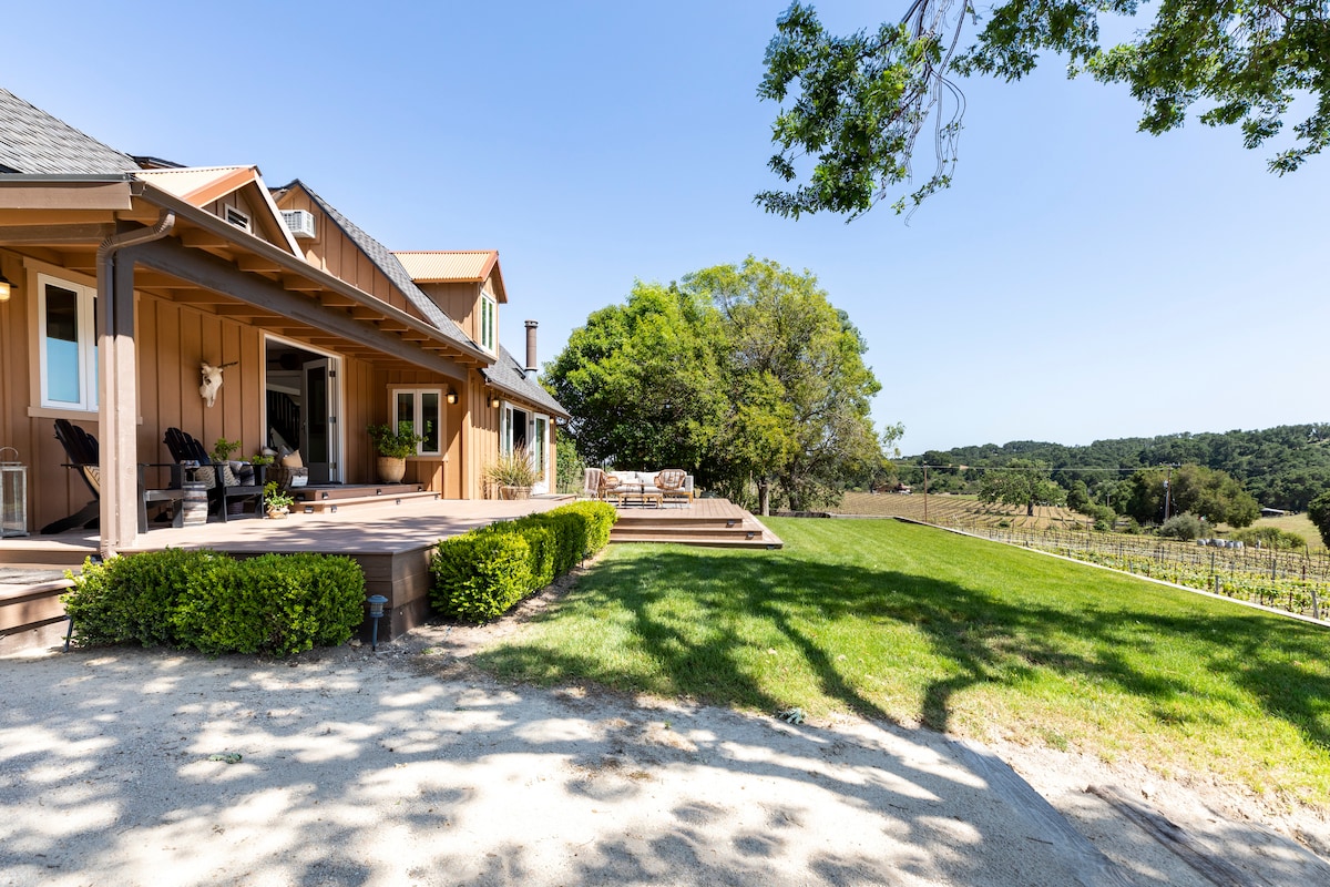 The exterior of the farmhouse is shown, featuring a spacious wooden deck with seating options. Lush greenery surrounds the home, leading to expansive vineyard views and rolling hills in the background. A shade tree offers a cool spot near the deck.