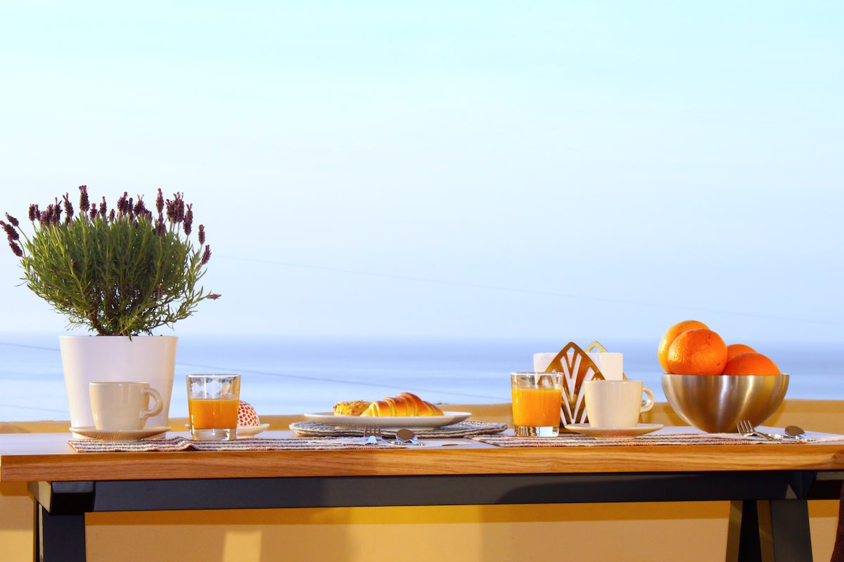 A dining table is set on a terrace, featuring a bowl of oranges, a basket with a pastry, and two glasses of orange juice. A potted plant adds a touch of greenery. The backdrop showcases a clear sea view, enhancing the inviting atmosphere.