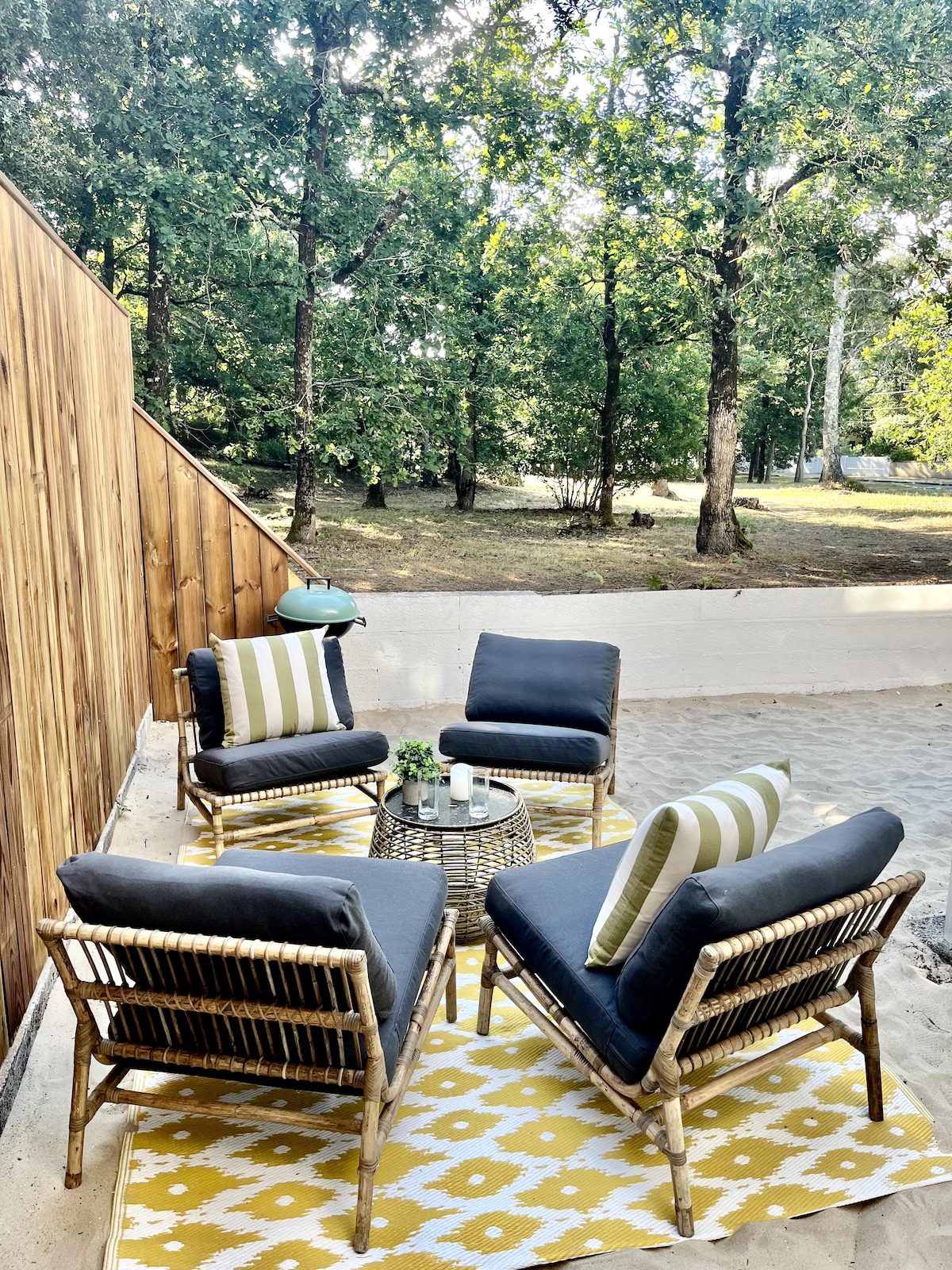 A private outdoor seating area contains four cushioned chairs arranged around a circular table. A patterned yellow rug adds warmth to the space, while trees provide a natural backdrop. A small grill is visible in the corner, creating an inviting atmosphere for relaxation.