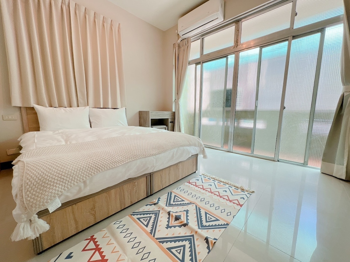 A spacious sleeping area features a comfortable bed dressed in neutral bedding, complemented by a textured throw. Large glass doors allow natural light to fill the room, leading to a balcony. Soft curtains frame the windows, creating a relaxed atmosphere.