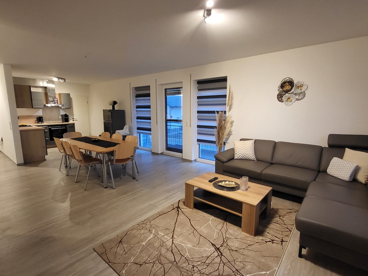 A modern living area features a spacious sectional sofa and a wooden coffee table atop an area rug. A dining table with six chairs is positioned nearby. Large windows with dual shades allow natural light to fill the room, enhancing the contemporary decor.