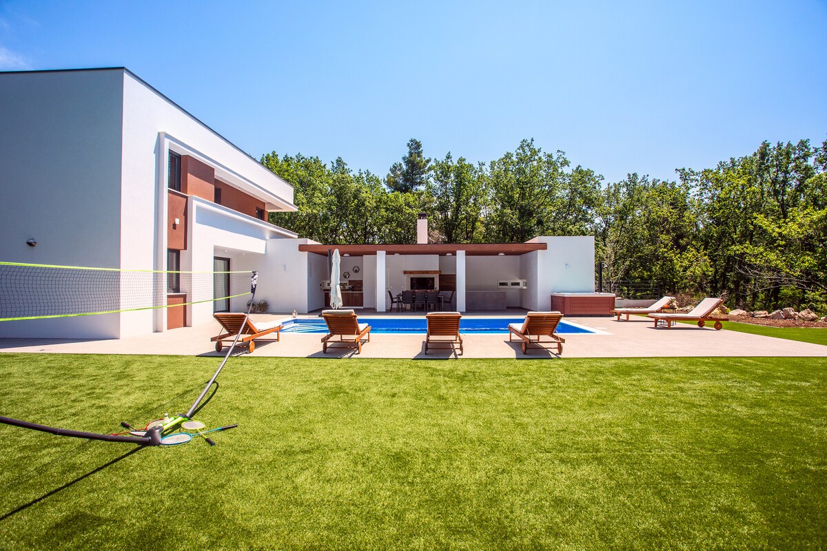 A private outdoor area features a heated pool surrounded by lounge chairs. A small badminton court is visible to the left on a well-maintained lawn. The villa's modern design is complemented by a shaded patio space in the background.