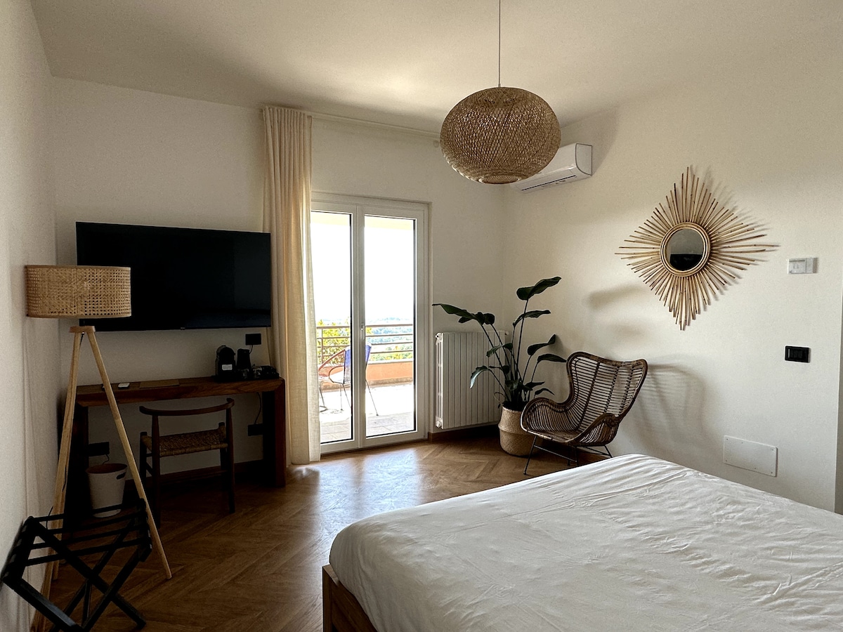 A bright and airy room features a spacious bed, a stylish rattan chair, and a wooden desk with a flat-screen television. Natural light floods in through glass doors that open to a balcony, with a potted plant adding a touch of greenery.