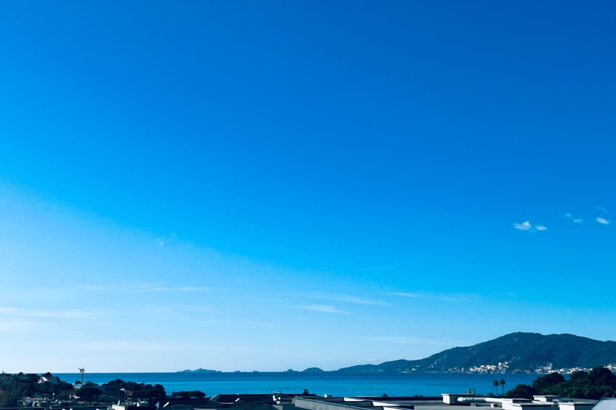 A panoramic view showcases a clear blue sky stretching over the tranquil sea, with distant islands visible on the horizon. The scene highlights the natural beauty of the surroundings, inviting relaxation and appreciation of the coastal landscape.