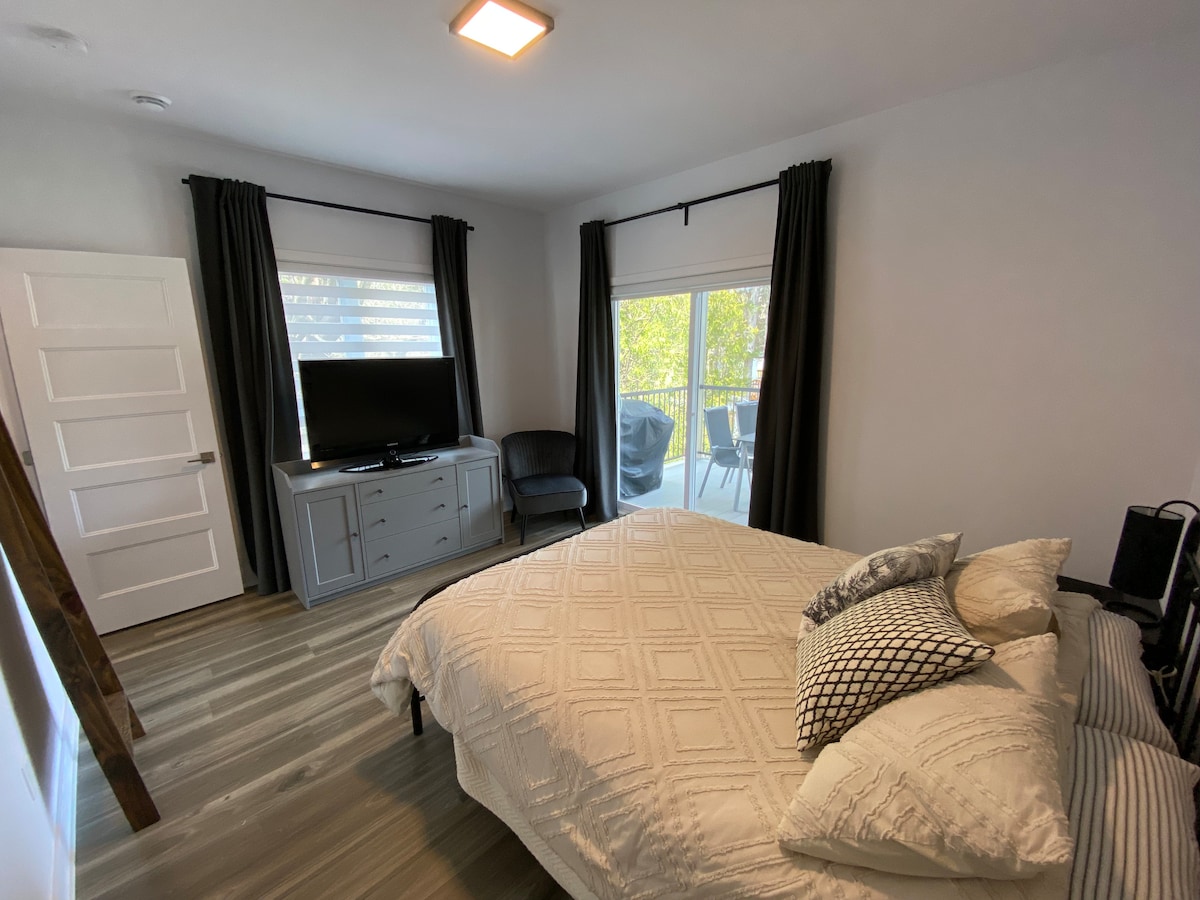 A cozy bedroom is presented, featuring a spacious bed adorned with patterned pillows. A television is mounted on a gray dresser, and a seating area is available with a chair. Natural light enters through glass doors leading to a terrace.