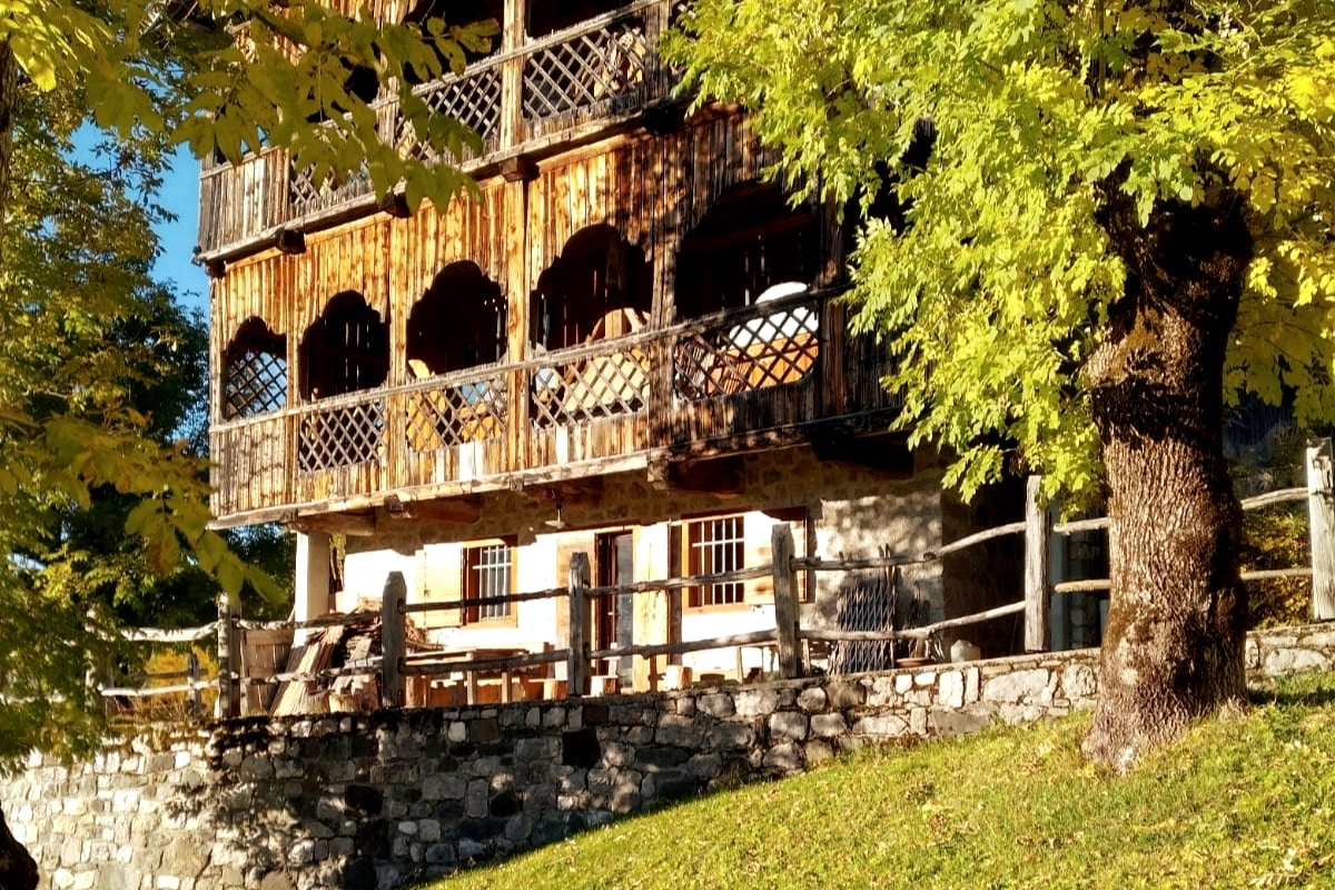 The rustic exterior of the historic building features wooden balconies and archway openings. Sunlight casts a warm glow on the thick green foliage surrounding the structure, while a stone wall and wooden fence define the outdoor area. A table is positioned on the lower level.