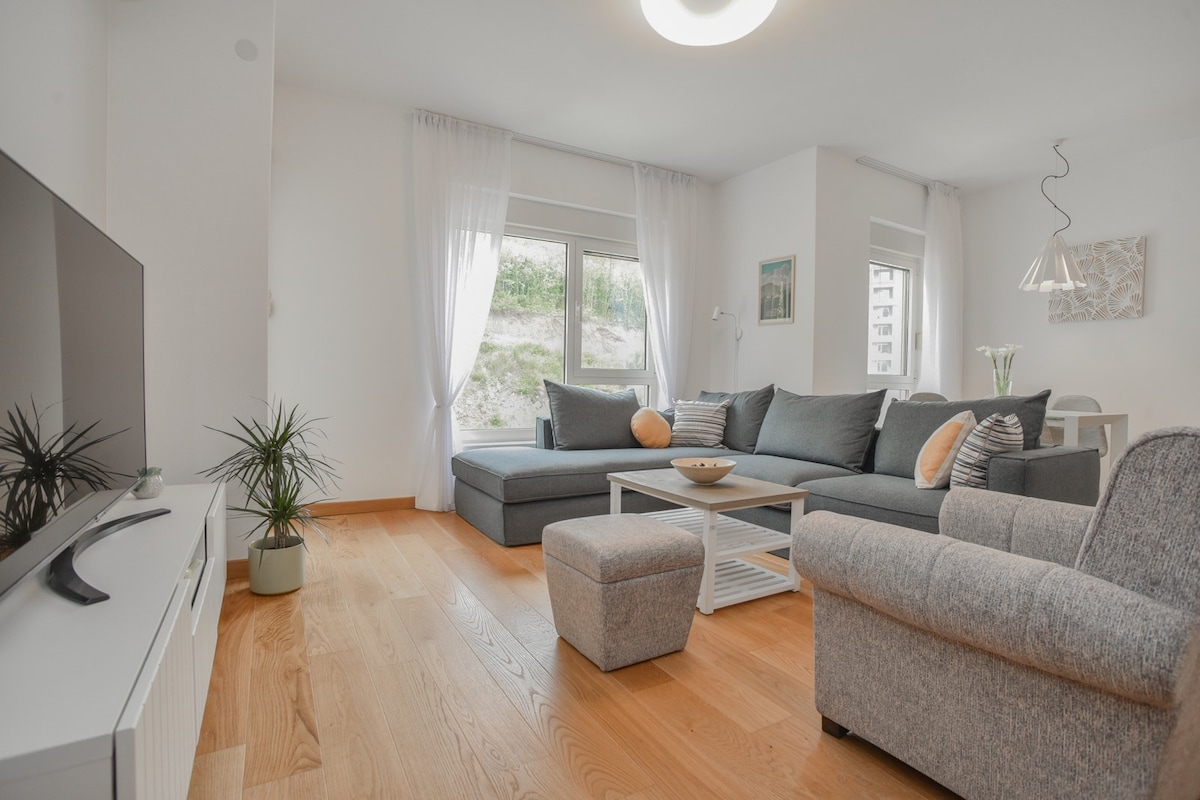 A modern living area is shown with a large sectional sofa and complementary armchair. Light-colored wood flooring extends throughout the space. A flat-screen TV is positioned on a minimalist media unit, and natural light enters through expansive windows, complemented by sheer white curtains.