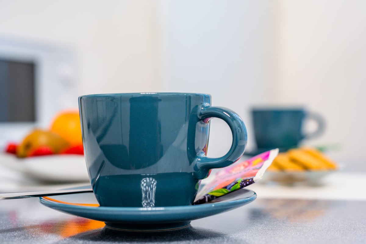 A teal coffee cup is positioned on a matching saucer, with a packet of sugar resting beside it. In the background, a plate with assorted fruits and biscuits is partially visible, contributing to a welcoming kitchen setting.