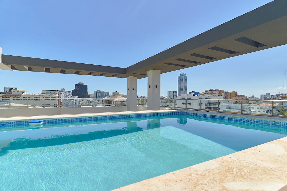 A spacious rooftop pool area is highlighted, featuring clear blue water and modern architectural lines. The surrounding terrace offers stunning views of the city skyline under a clear blue sky, with ample space for relaxation and sunbathing.
