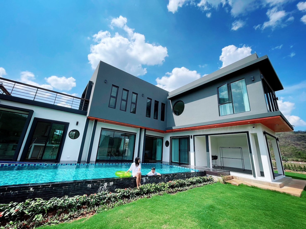A modern villa is set against a clear blue sky, featuring a spacious pool surrounded by lush green grass and well-maintained landscaping. Large windows reflect the natural light, highlighting the sleek architectural design and open layout of the facility.