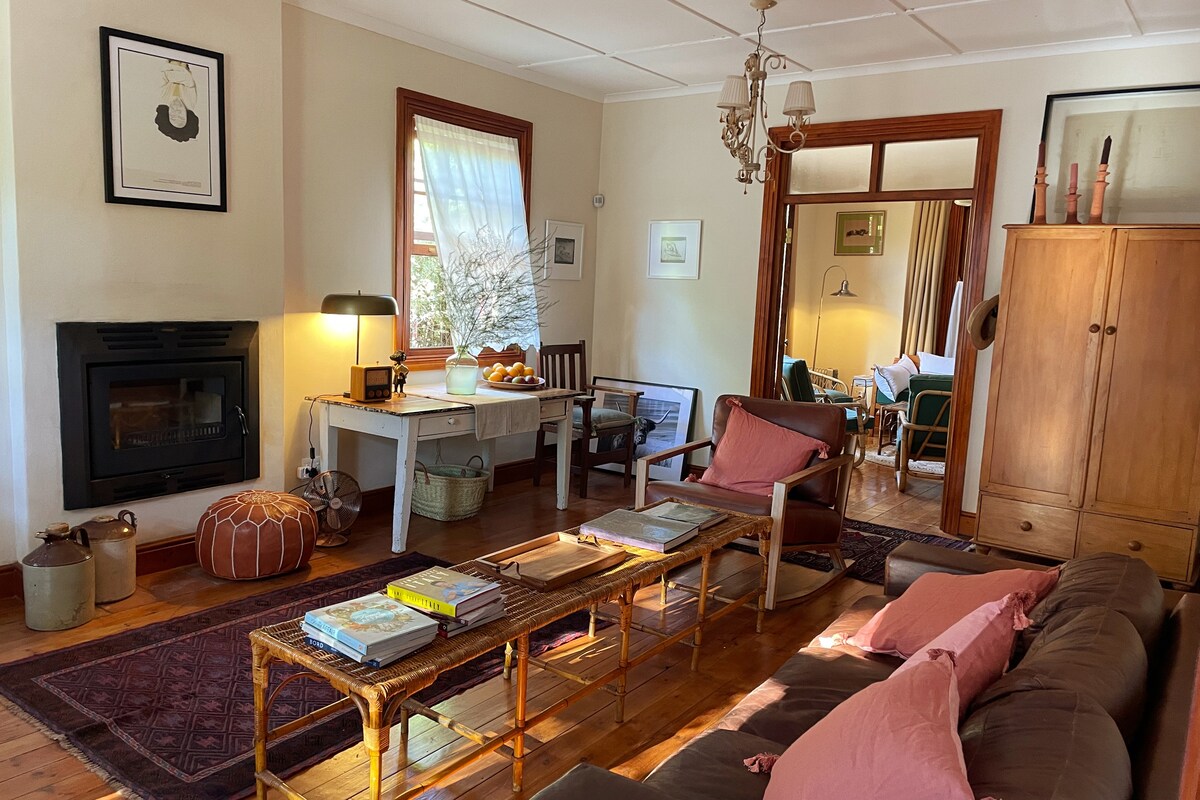 The inviting living area features a comfortable sofa adorned with pink cushions. A wooden table is set near a cozy fireplace, while natural light enters through large windows. A woven pouf and wooden storage unit add to the warmth, with books and magazines displayed on the coffee table.