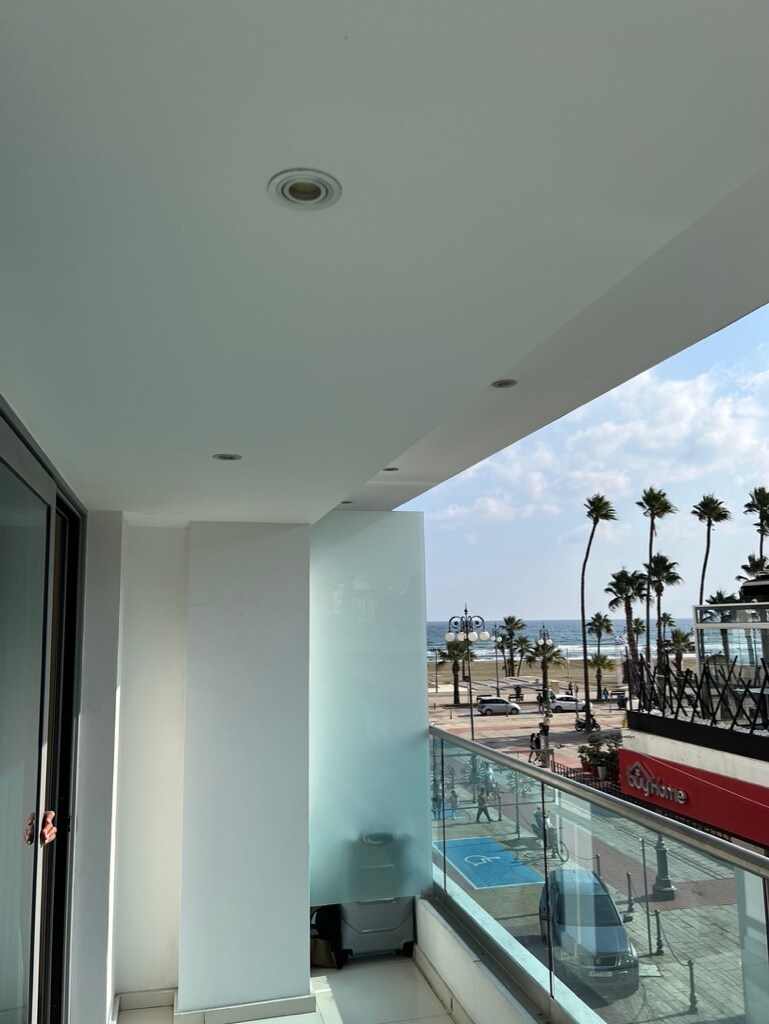 A balcony view captures the sea in the distance, bordered by palm trees lining the promenade below. The space is illuminated by natural light, with a minimalist design featuring smooth white walls and a glass railing, enhancing the open atmosphere.