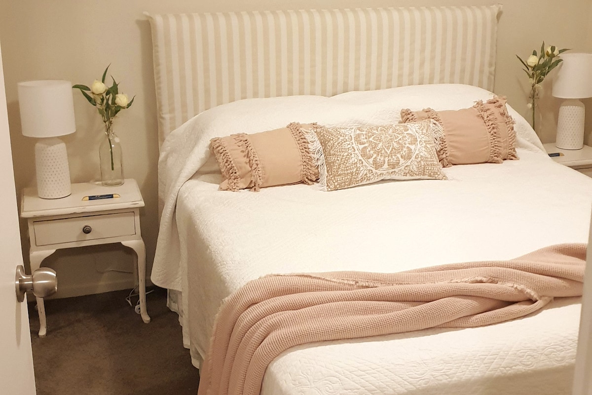 A cozy bedroom is showcased with a spacious bed draped in light-colored bedding. Decorative pillows and a soft throw blanket add subtle warmth. Each side of the bed is complemented by bedside tables with lamps, while fresh flowers enhance the serene environment.