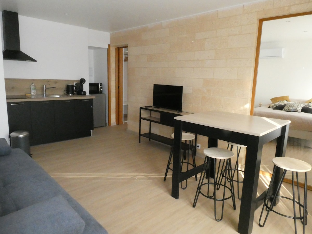 An open living space features light-colored walls and a wooden floor. A kitchenette is equipped with modern appliances, including a coffee machine. A dining table with high stools sits centrally, while a television on a low stand faces a comfortable sofa. A partially visible bedroom is located through an adjoining door.