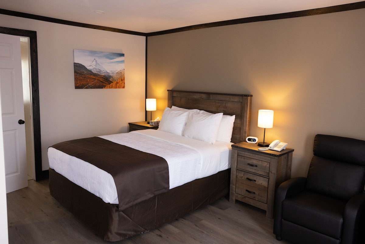 A cozy bedroom features a spacious bed dressed in white linens and a brown bed skirt. Two bedside lamps provide soft illumination, while a wooden nightstand and a reclining chair enhance the space. A mountain landscape adorns the wall above the bed.