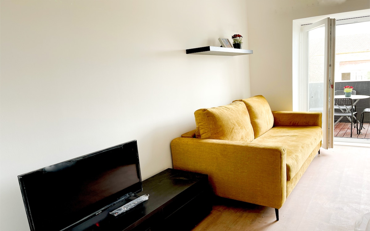 A modern living area features a vibrant yellow sofa, positioned against a clean white wall. A dark media console is located beneath a flat-screen television. Light enters the space through a door that opens to a small outdoor seating area with a dining set.