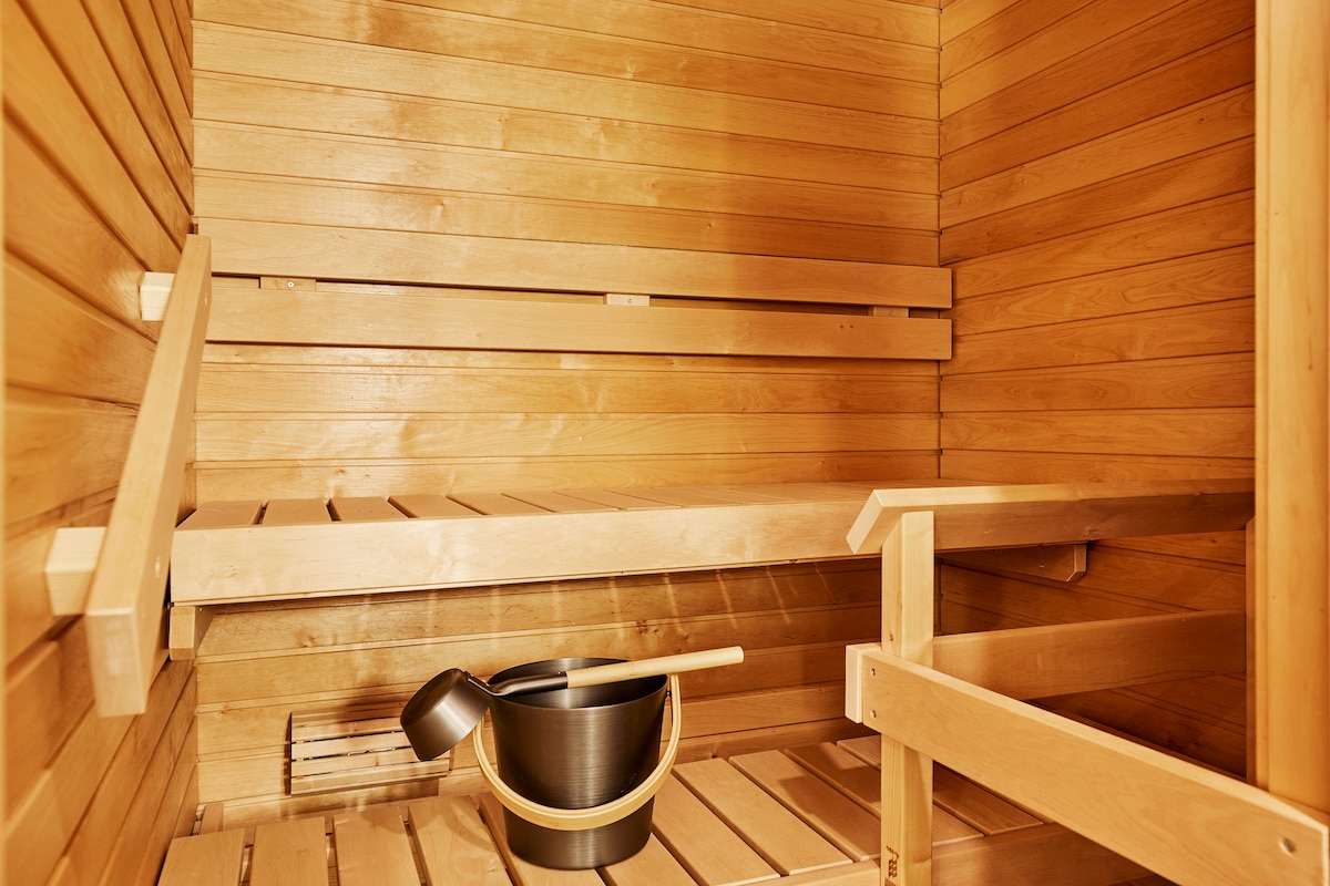 A wooden sauna interior presents smooth, warm-toned walls and benches made of light timber. A metal bucket and ladle sit on a shelf, adding to the traditional sauna atmosphere. The space is designed for relaxation and wellness.
