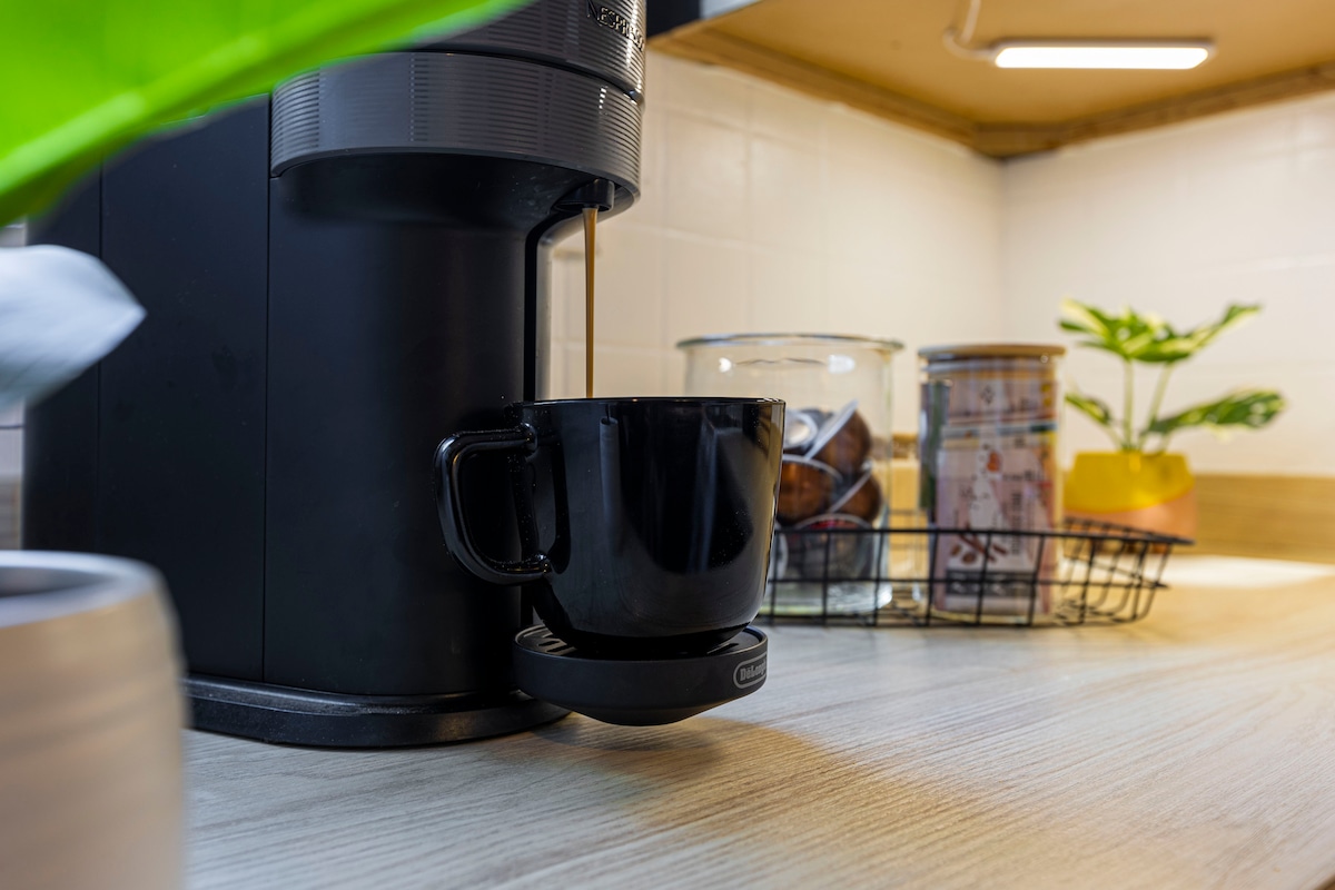 A coffee machine sits on a wooden countertop, pouring a fresh cup of coffee into a black mug. Various jars containing coffee pods and a small potted plant are positioned nearby, contributing to the kitchen's functionality.