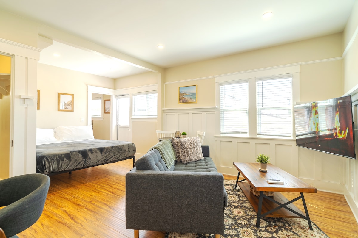 The studio features a spacious layout with a comfortable sofa facing a mounted flat screen television. Natural light fills the room through multiple windows. A king-sized bed is positioned in the background, and a wooden coffee table adds a cozy touch to the living area.