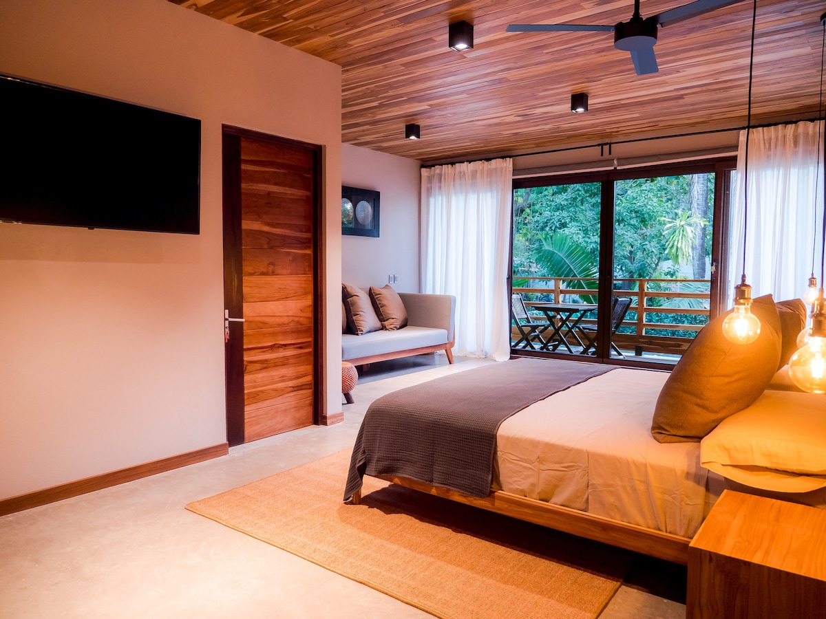 A serene bedroom showcases a spacious bed with plush pillows, accompanied by bedside tables with stylish lamps. Large glass doors provide views of the lush surroundings, while subtle wooden accents create a calm atmosphere. A seating area is positioned near the window, enhancing the inviting space.