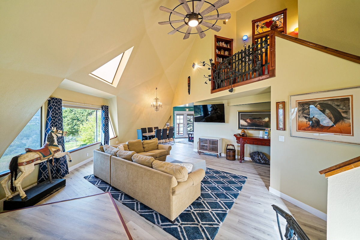 A spacious living area features high, angled ceilings and large windows that invite natural light. A comfortable beige sectional sofa is positioned on a patterned rug, while a decorative horse sculpture adds character. A balcony overlooking the room is adorned with bookshelves and artwork.