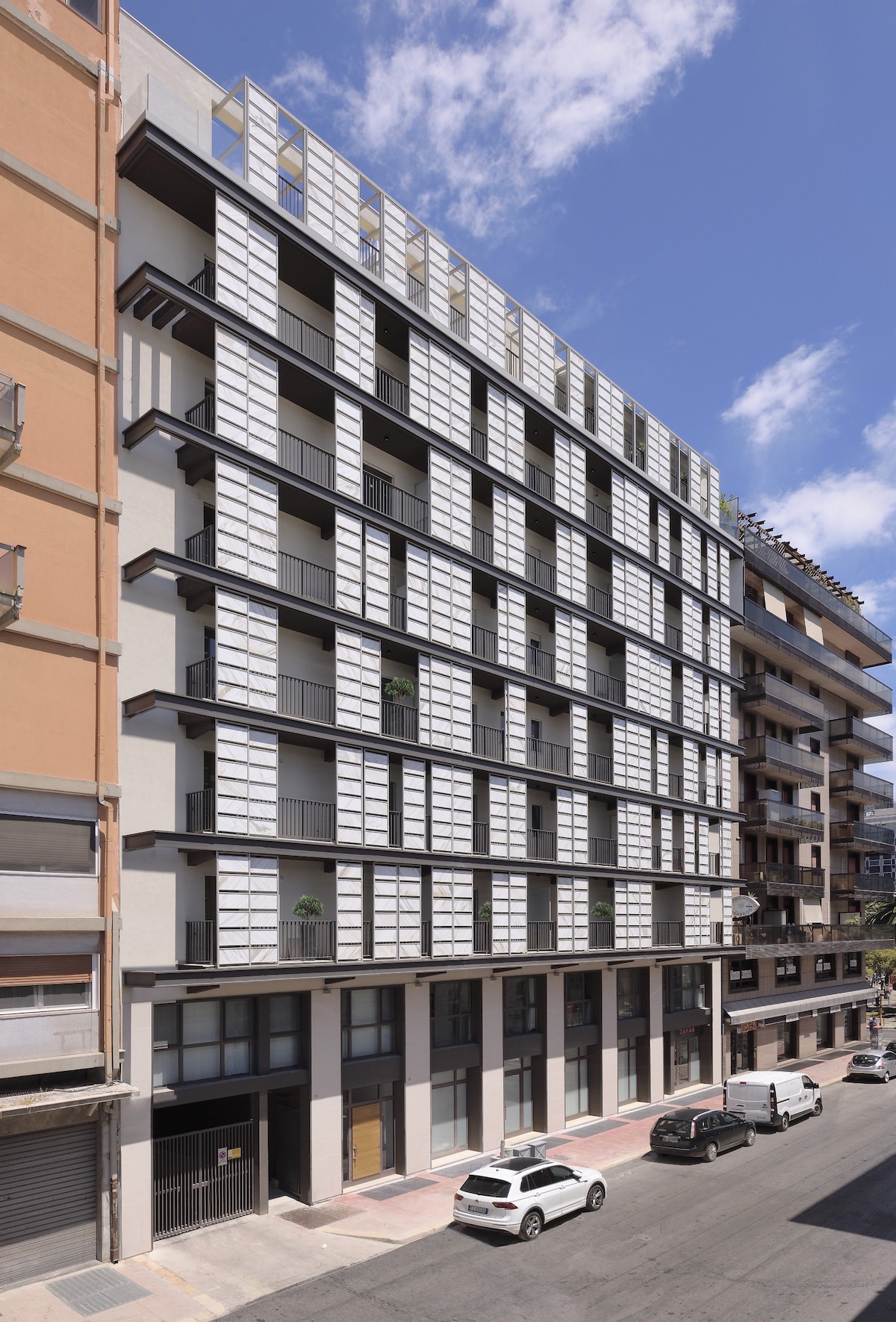 The modern building features a clean, symmetrical facade with expansive windows and distinctive horizontal elements. The structure includes multiple balconies, allowing for natural light, while the surrounding area showcases a mix of urban architecture, including both contemporary and older designs.